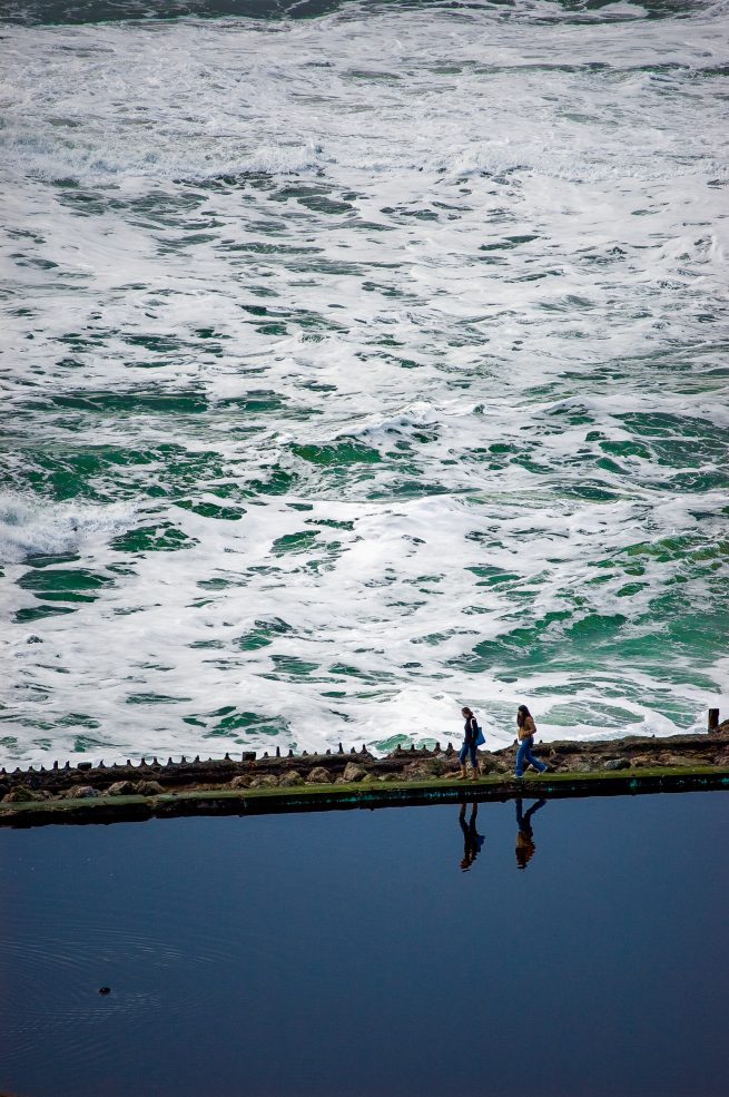 Two women walking along ocean, Sutro Baths ruins, San Francisco